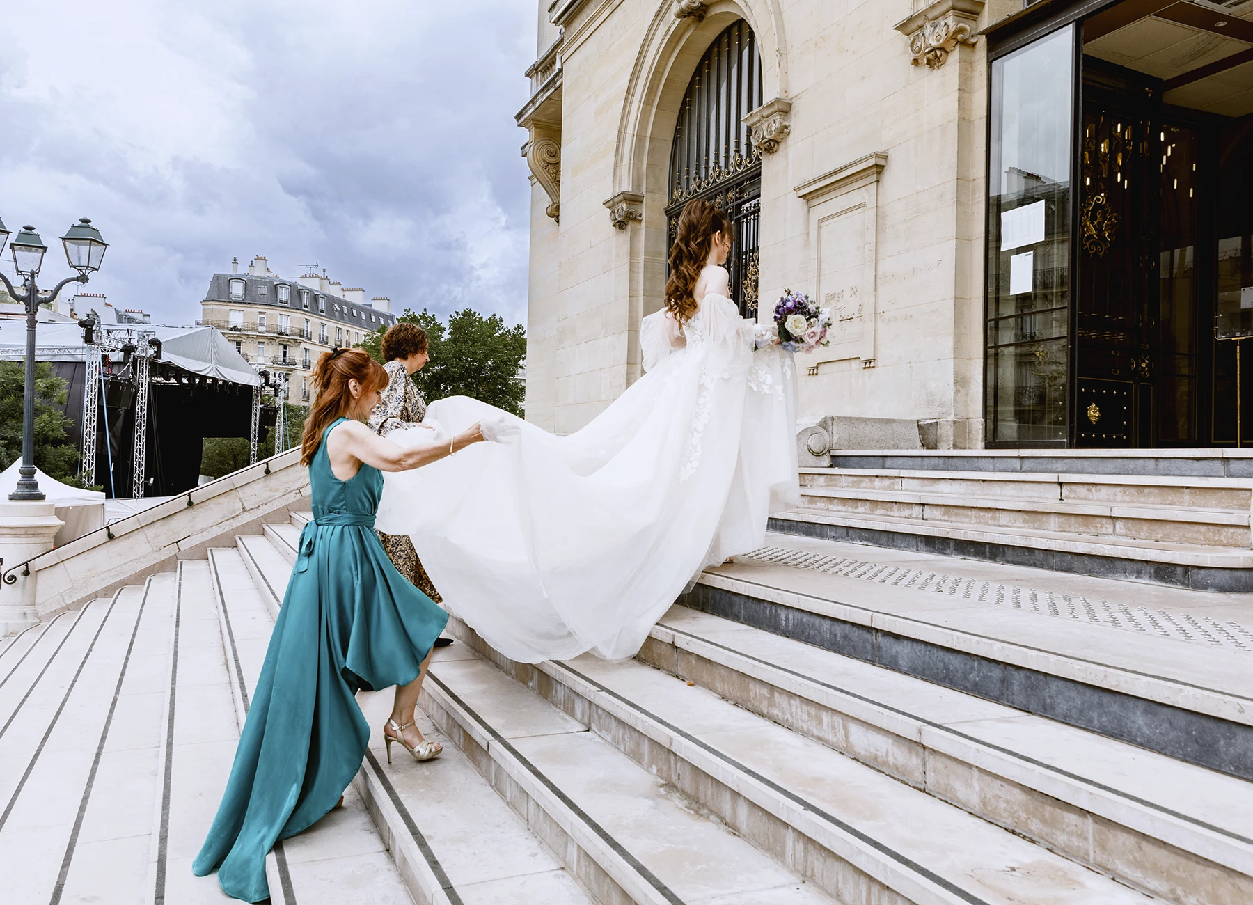 Mère de la mariée et une amie tenant la traîne pendant qu'elle monte les marches. Photo par Laurène Zabary.