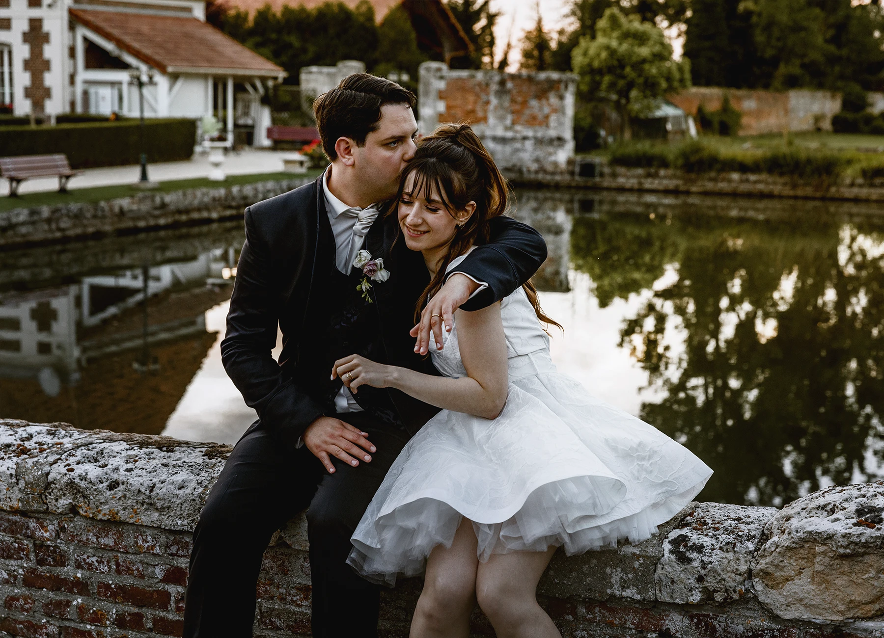 Marié embrassant la mariée sur la tête, assis sur une margelle au château de Quesmy, golden hour, photographie de Laurène Zabary
