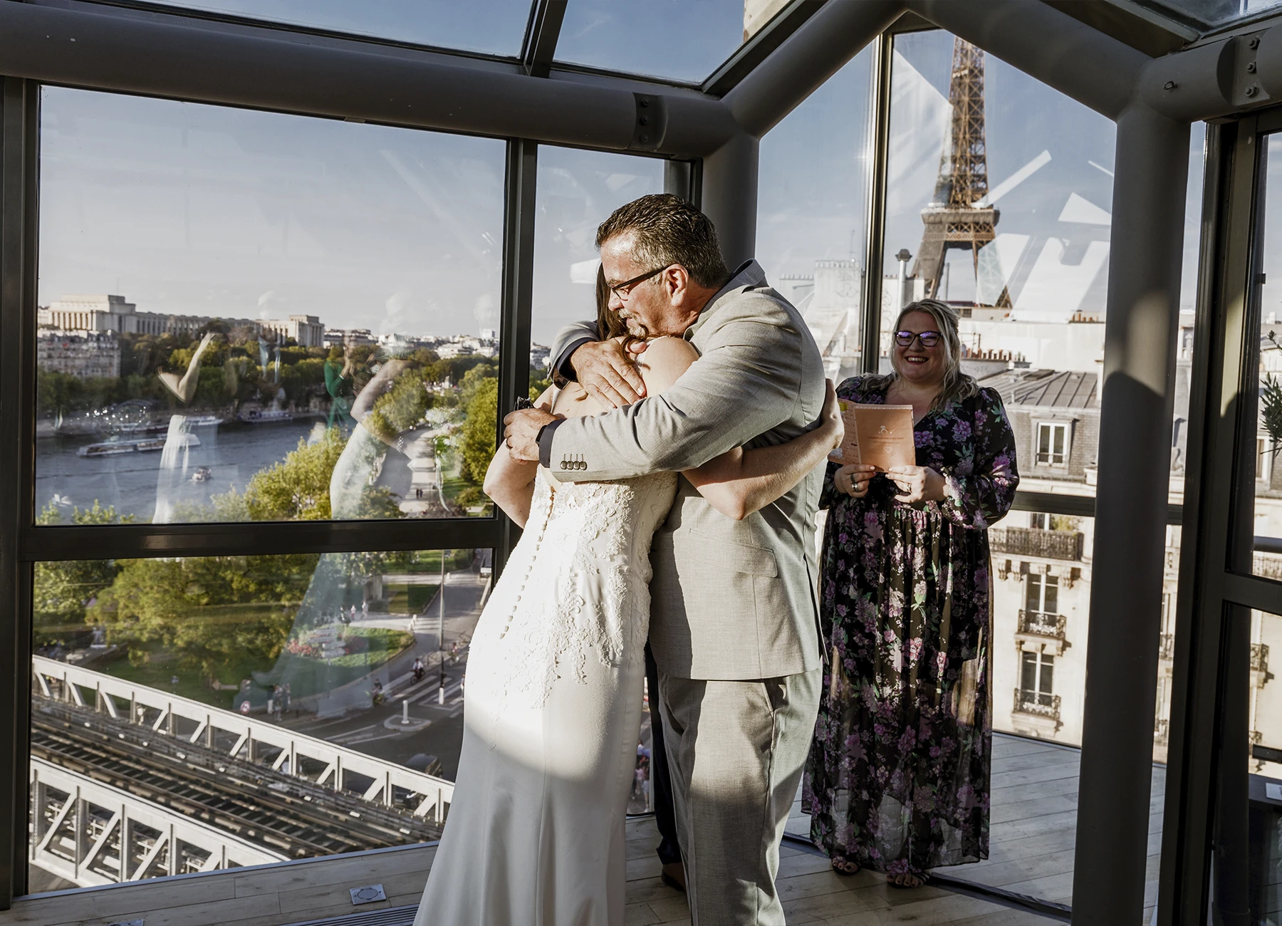 Mariée en robe blanche et son père en costume gris se serrant dans les bras avec la tour Eiffel en arrière-plan, capturés au Rooftop Grenelle à Paris par Laurène Zabary.