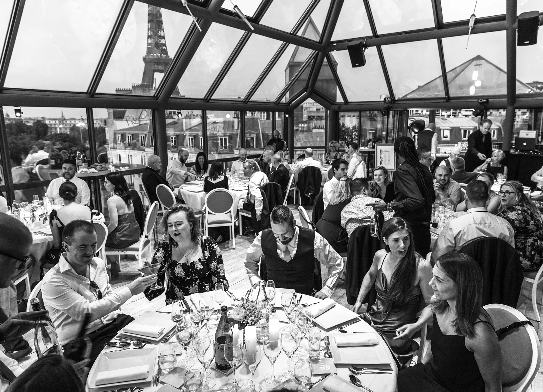 Vue large de la salle de réception sous la verrière pendant le repas de mariage au Rooftop Grenelle à Paris, capturée en noir et blanc par Laurène Zabary.