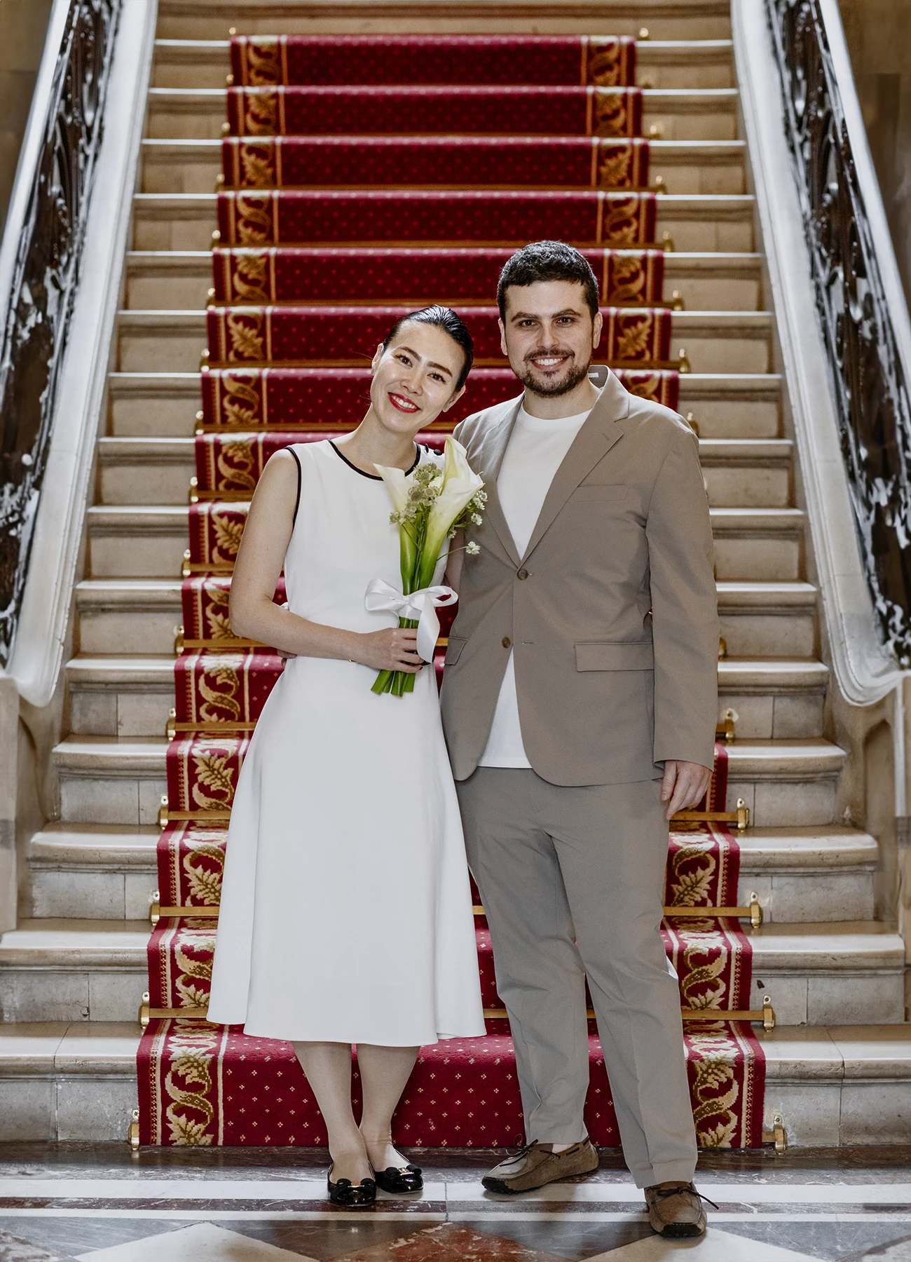 Couple de mariés devant les escaliers de la mairie du 7e arrondissement de Paris, la mariée en robe blanche avec un bouquet de fleurs.