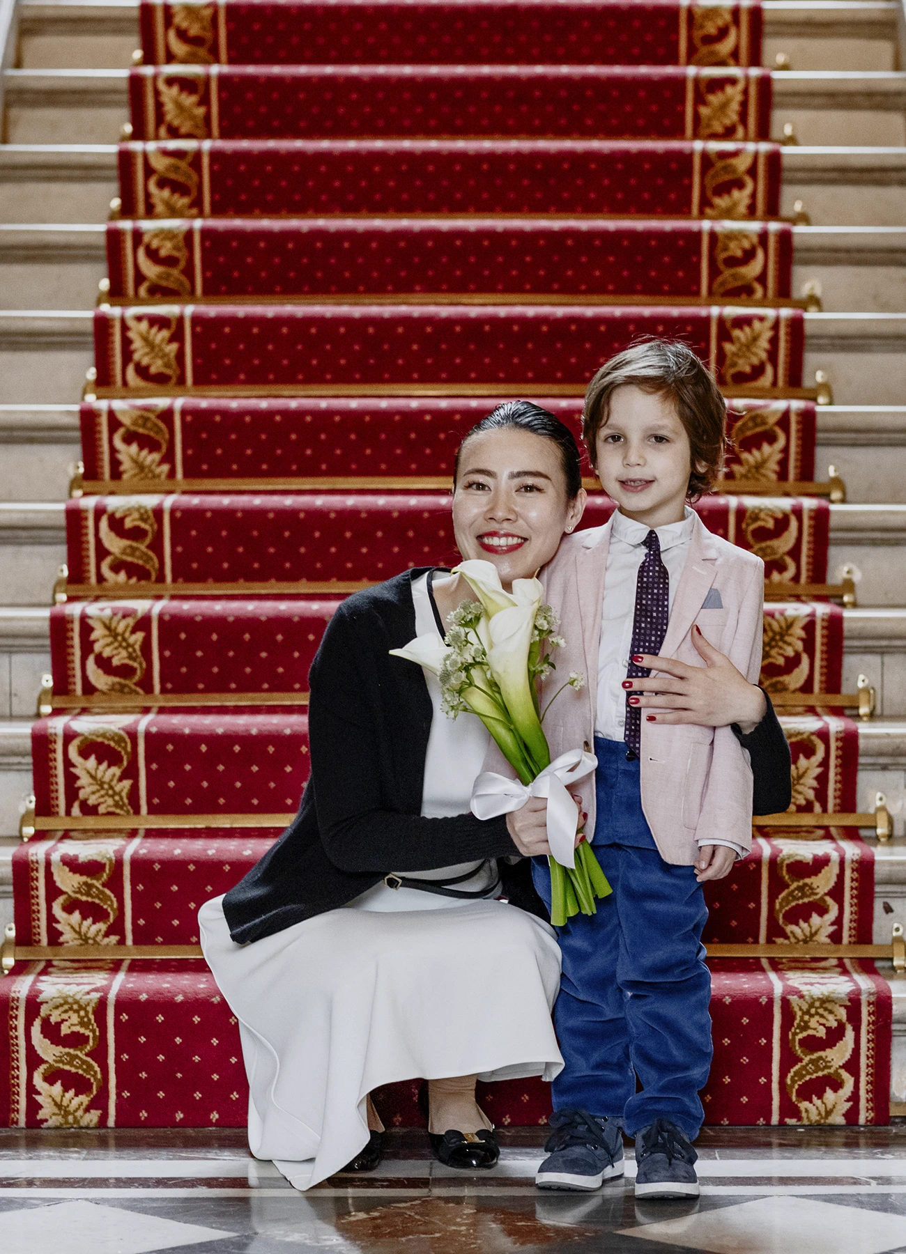 La mariée en robe blanche avec un bouquet de lys Calla, accroupie à côté d'un jeune garçon devant les escaliers de la mairie du 7e arrondissement