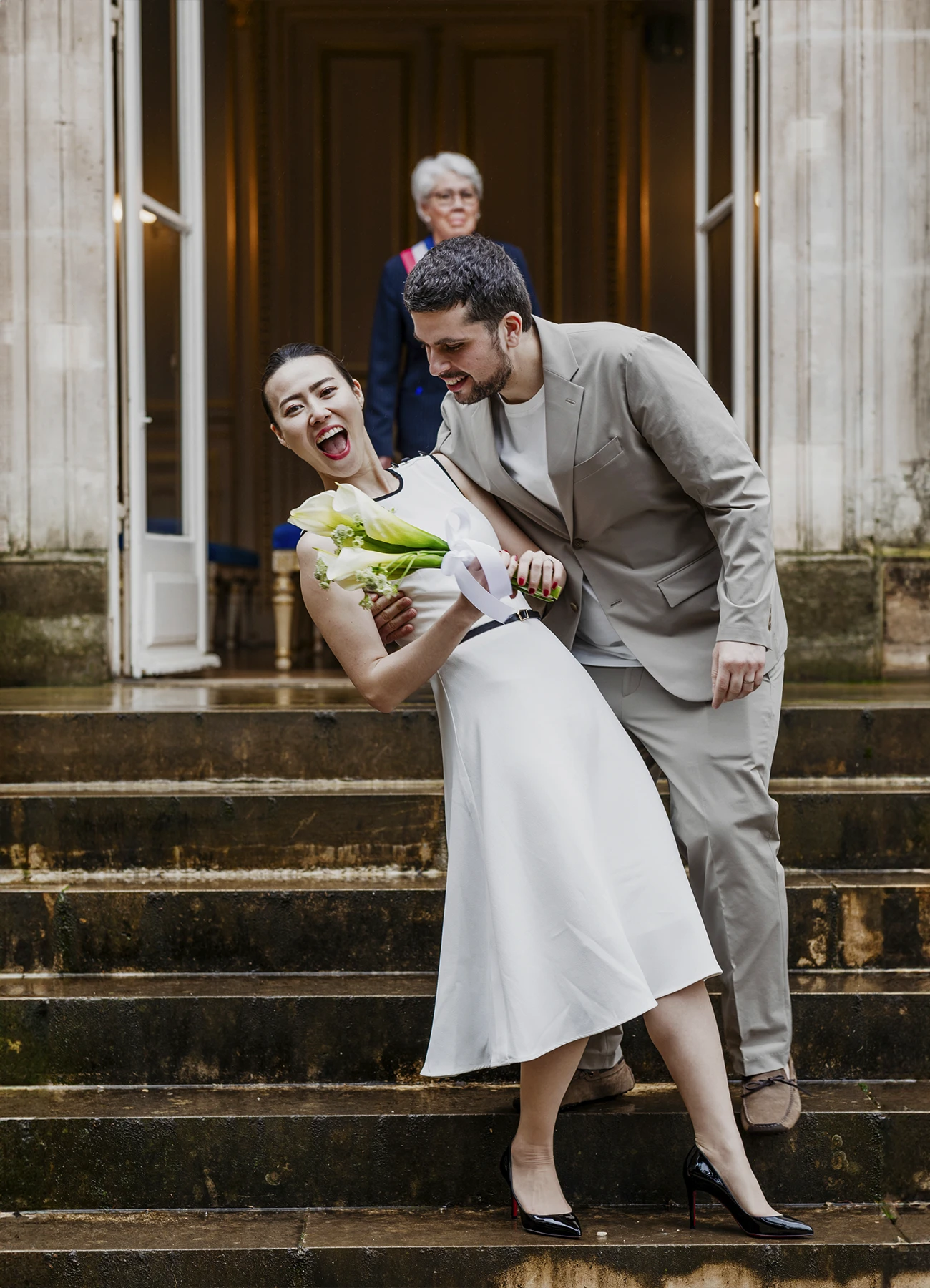 Le marié tient sa femme dans les bras sur les marches de la mairie du 7e arrondissement de Paris, avec la mariée souriante à la caméra.