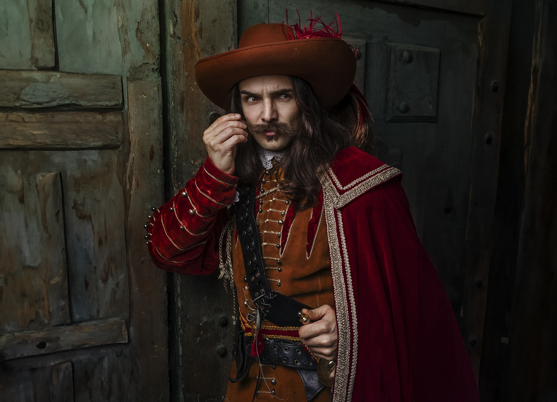 Comédien en costume de mousquetaire rouge avec moustaches et cheveux longs, posant contre une vieille porte en bois à Paris. Photo par Laurène Zabary.