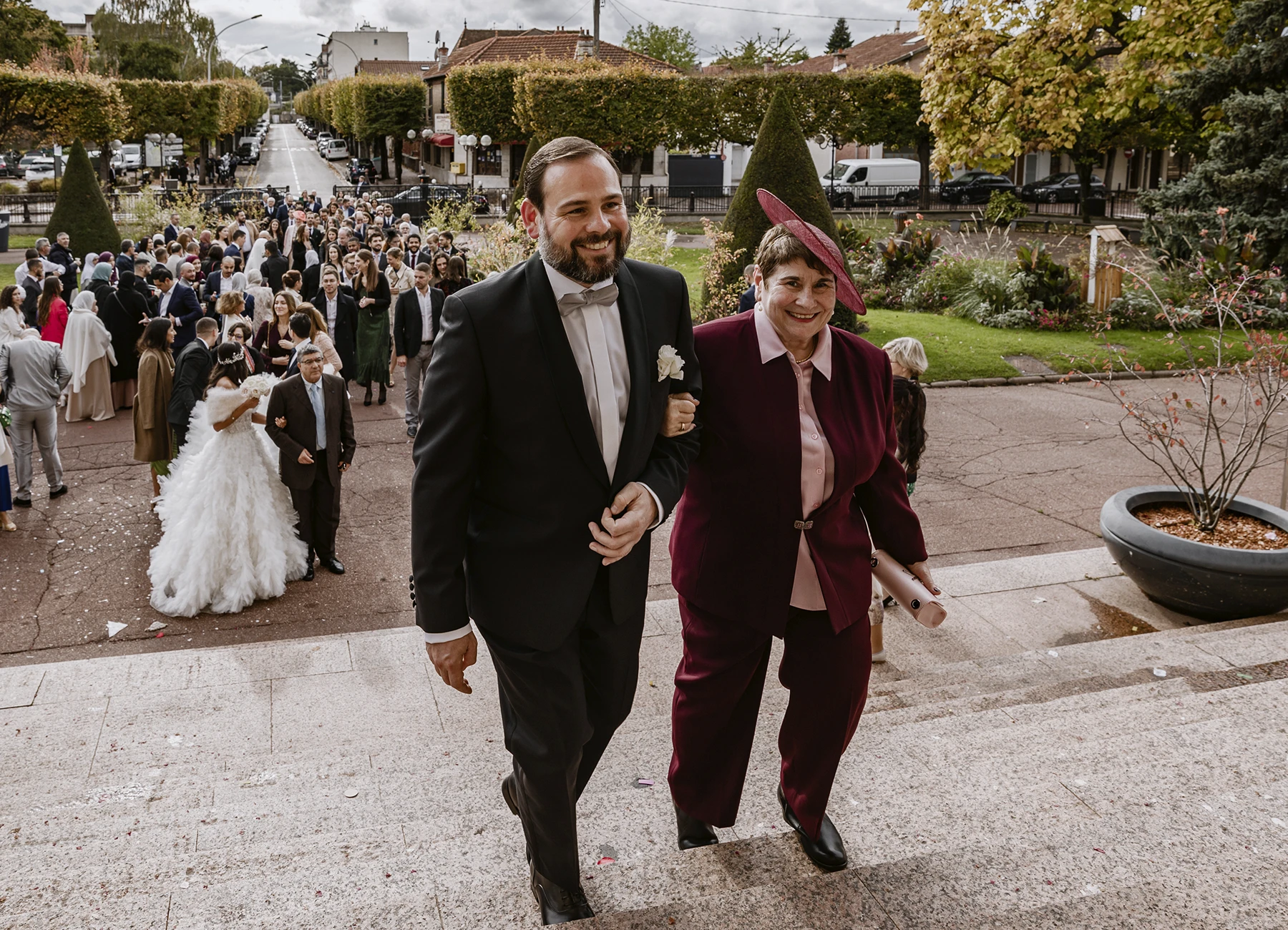 Marié entrant à la mairie au bras de sa mère habillée en rose/bordeaux avec chapeau assorti. Photo par Laurène Zabary