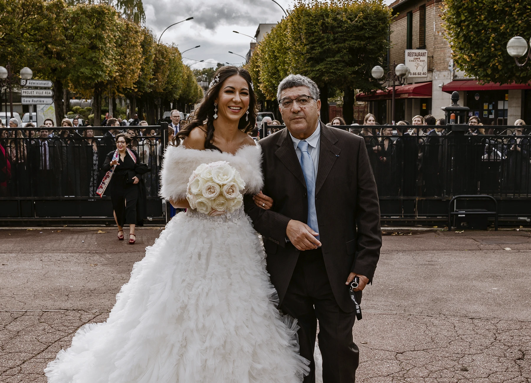 Mariée en robe blanche au bras de son père à l'entrée de la mairie, souriant à l'objectif. Photo par Laurène Zabary