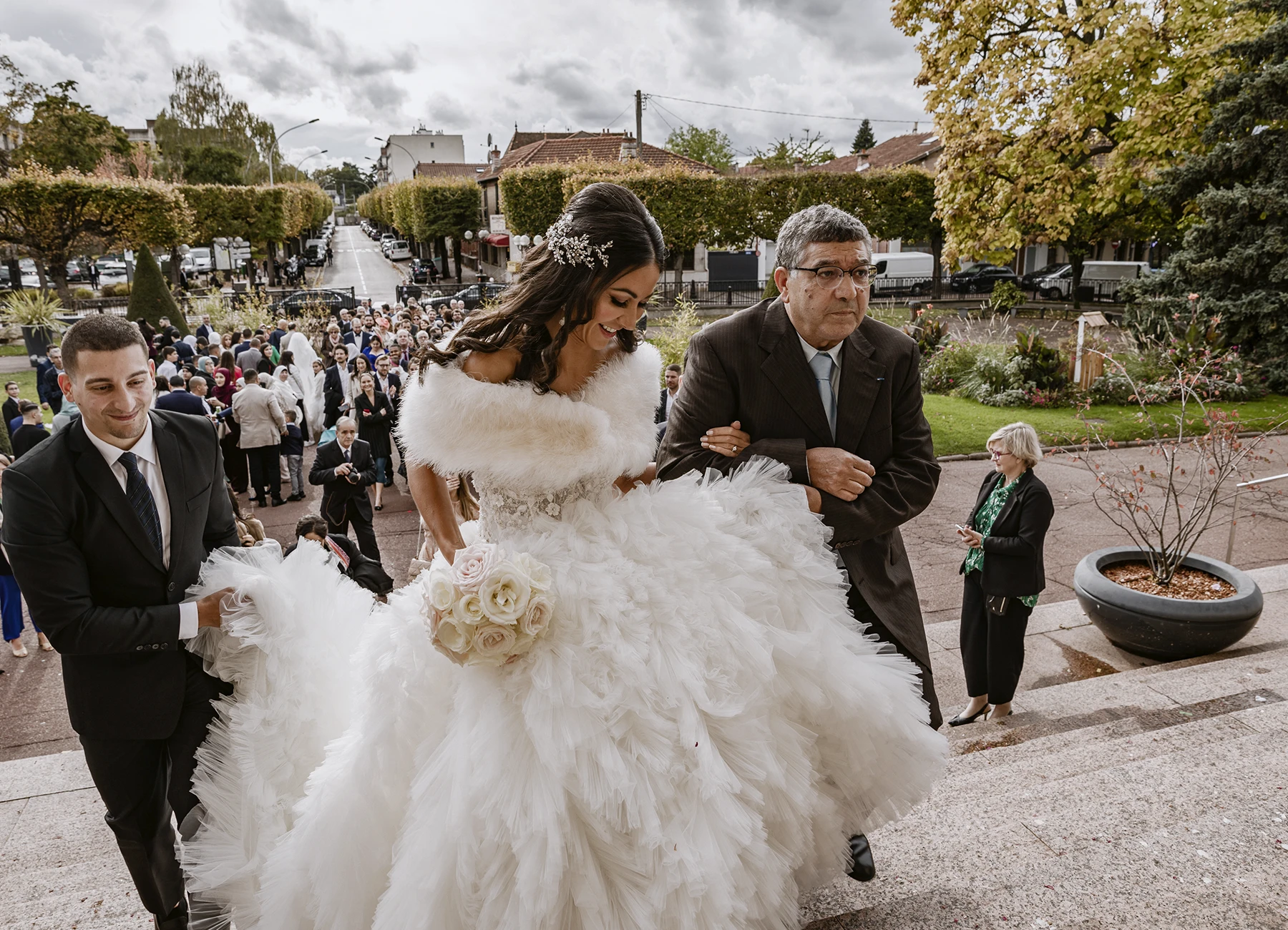 Mariée en robe blanche au bras de son père à l'entrée de la mairie, souriant à l'objectif. Photo par Laurène Zabary