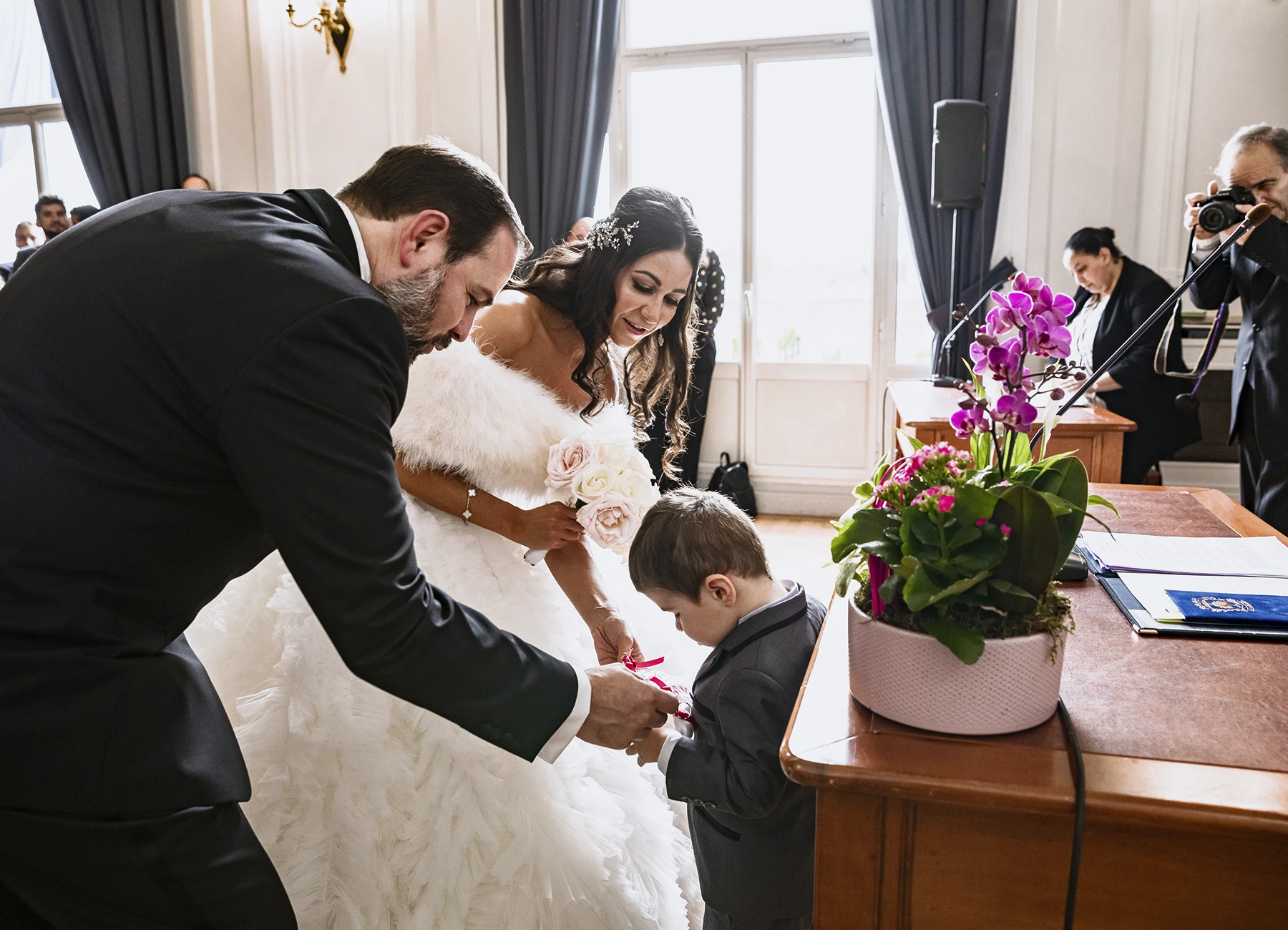 Mariés penchés vers un petit garçon présentant les alliances. Photo par Laurène Zabary