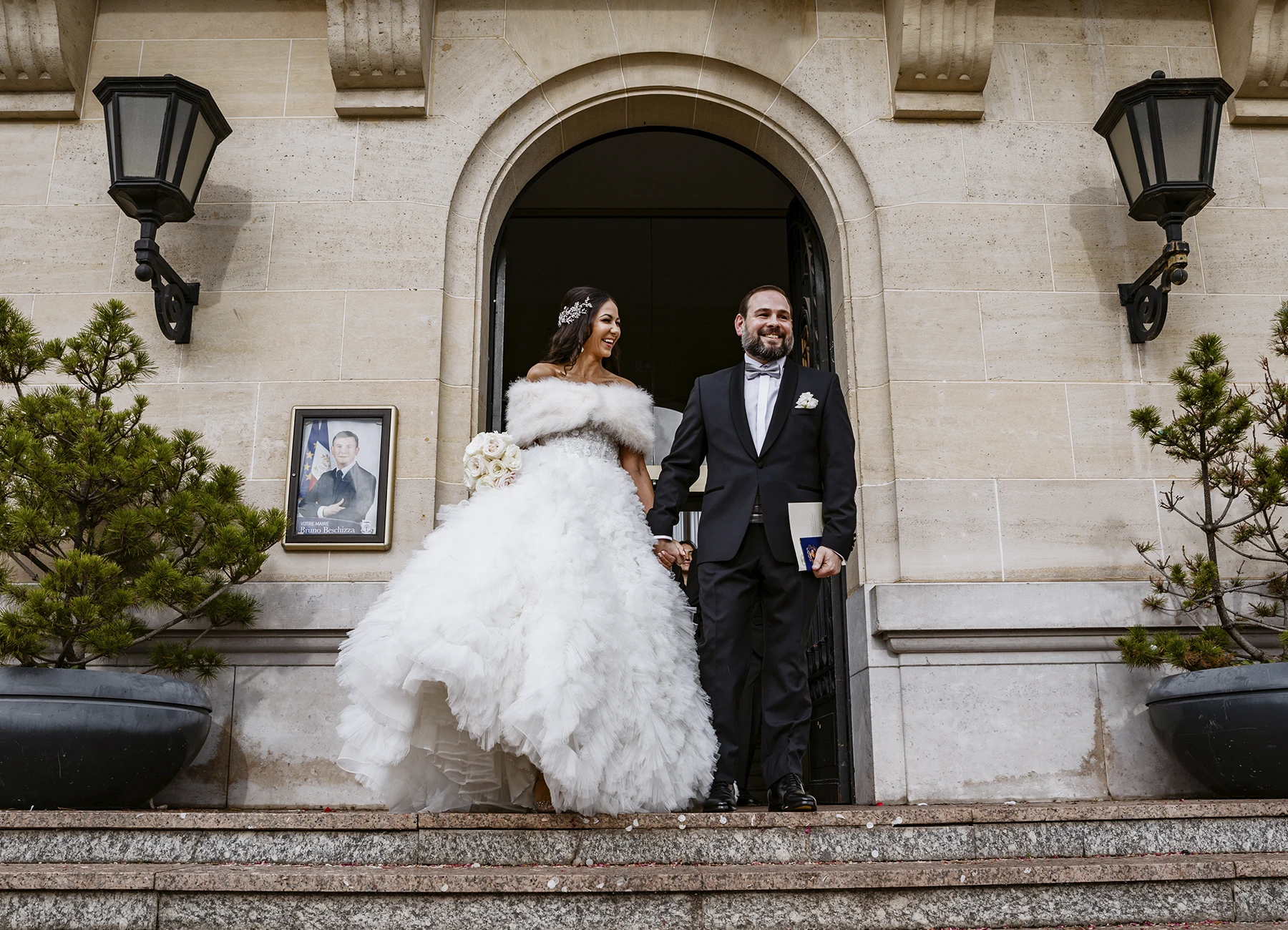 Les mariés souriant, se tenant la main à la porte de la mairie. La mariée tient son bouquet. Photo par Laurène Zabary