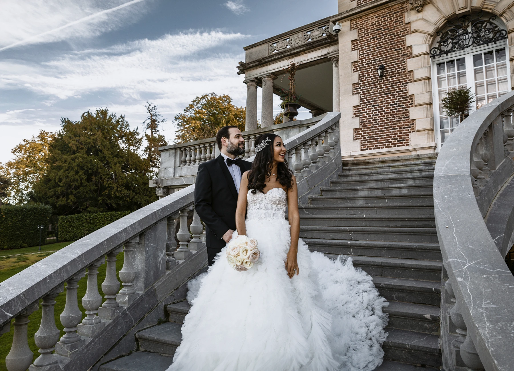 "Couple de jeunes mariés élégants sur les marches extérieures du Château Bouffémont, souriant et regardant sur leur gauche sous un magnifique ciel bleu. Photo par Laurène Zabary.