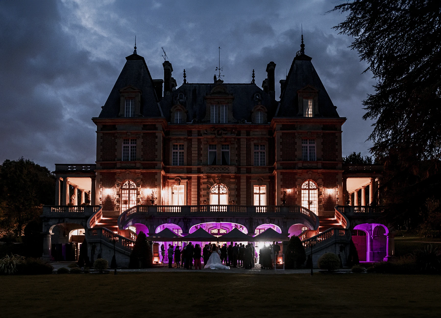 Château Bouffémont illuminé de nuit avec des éclairages nocturnes violet, jaune et orange, silhouettes des invités en arrière-plan. Photo par Laurène Zabary
