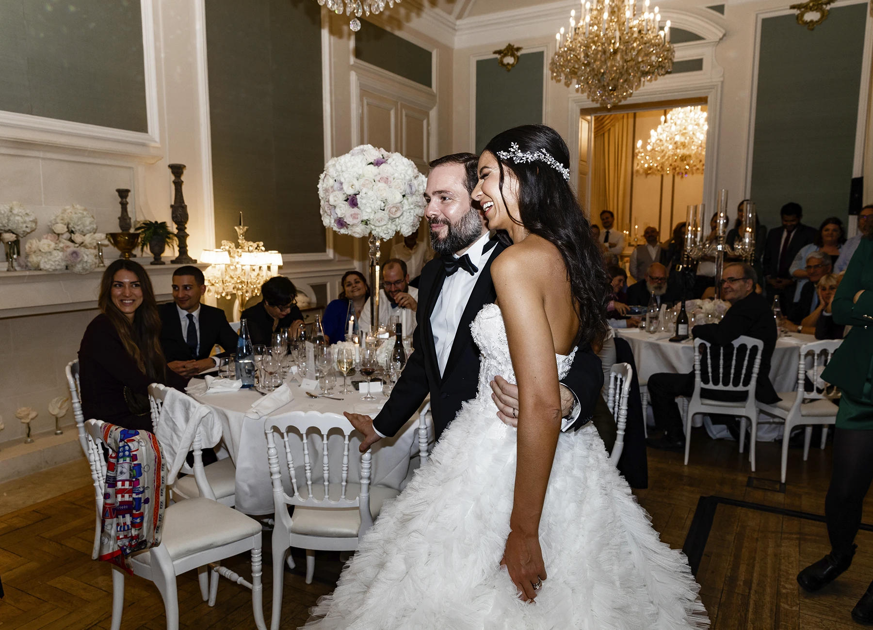Les mariés souriant à leurs invités dans la salle de réception du Château Bouffémont, tables décorées en arrière-plan. Photo par Laurène Zabary.