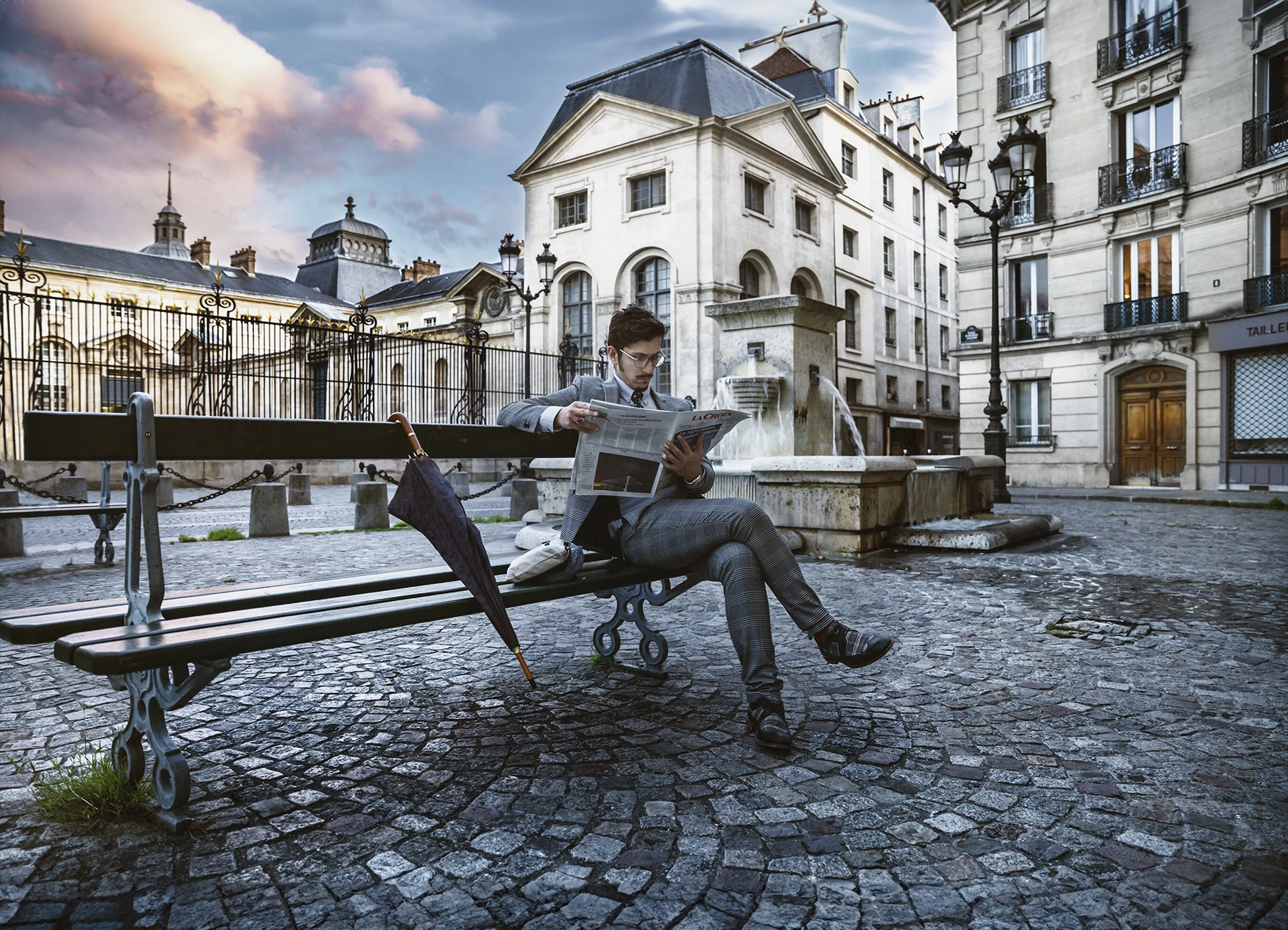Amir, homme en costume gris rayé, portant des lunettes, assis sur un banc devant le Val de Grace, lisant La Croix avec un parapluie posé à ses côté. Photo par Laurène Zabary.