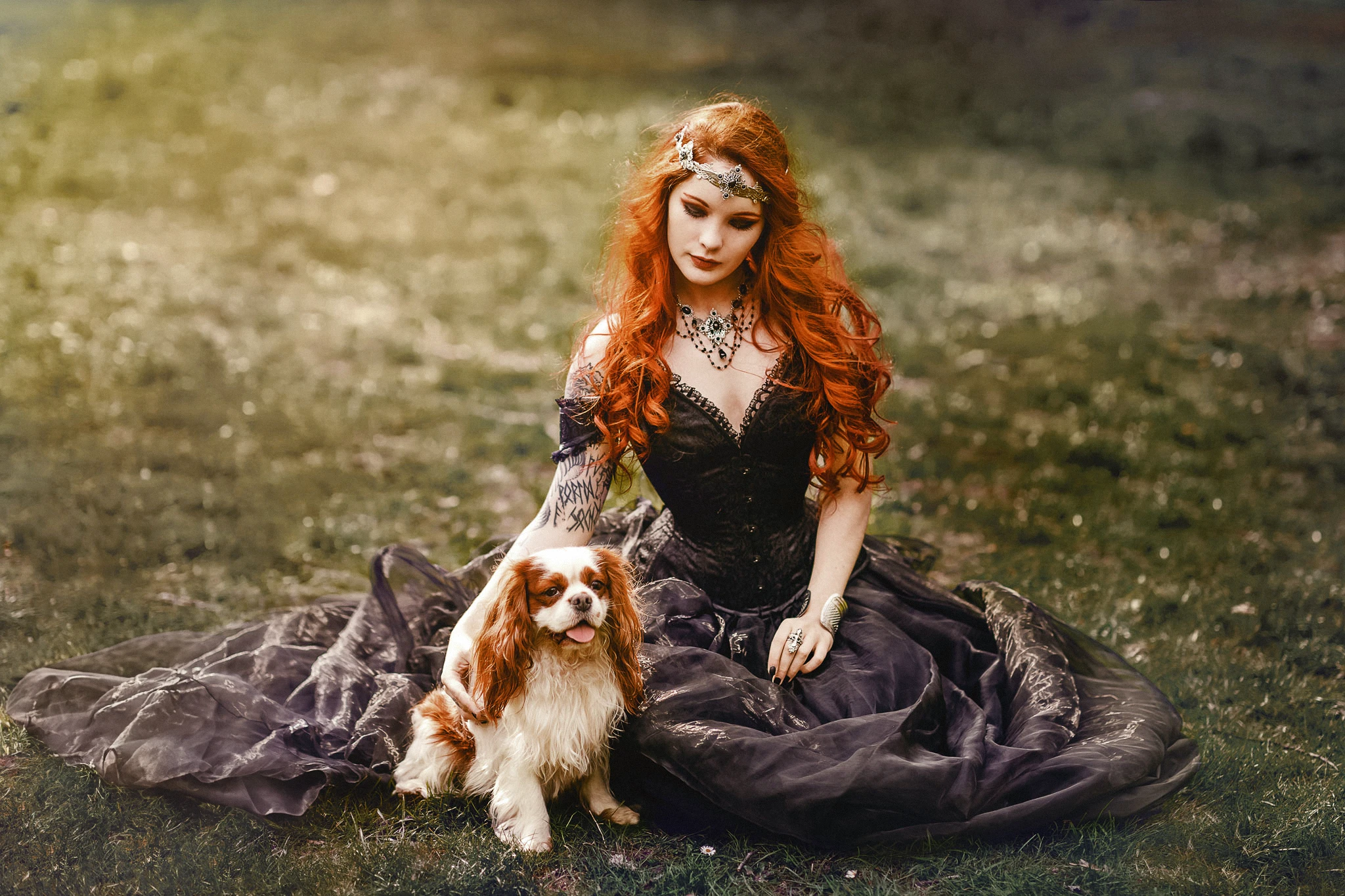 Elisa, jeune femme rousse tatouée, en robe de princesse noire, assise dans l'herbe caressant un chien Cavalier King Charles. Photo par Laurène Zabary.