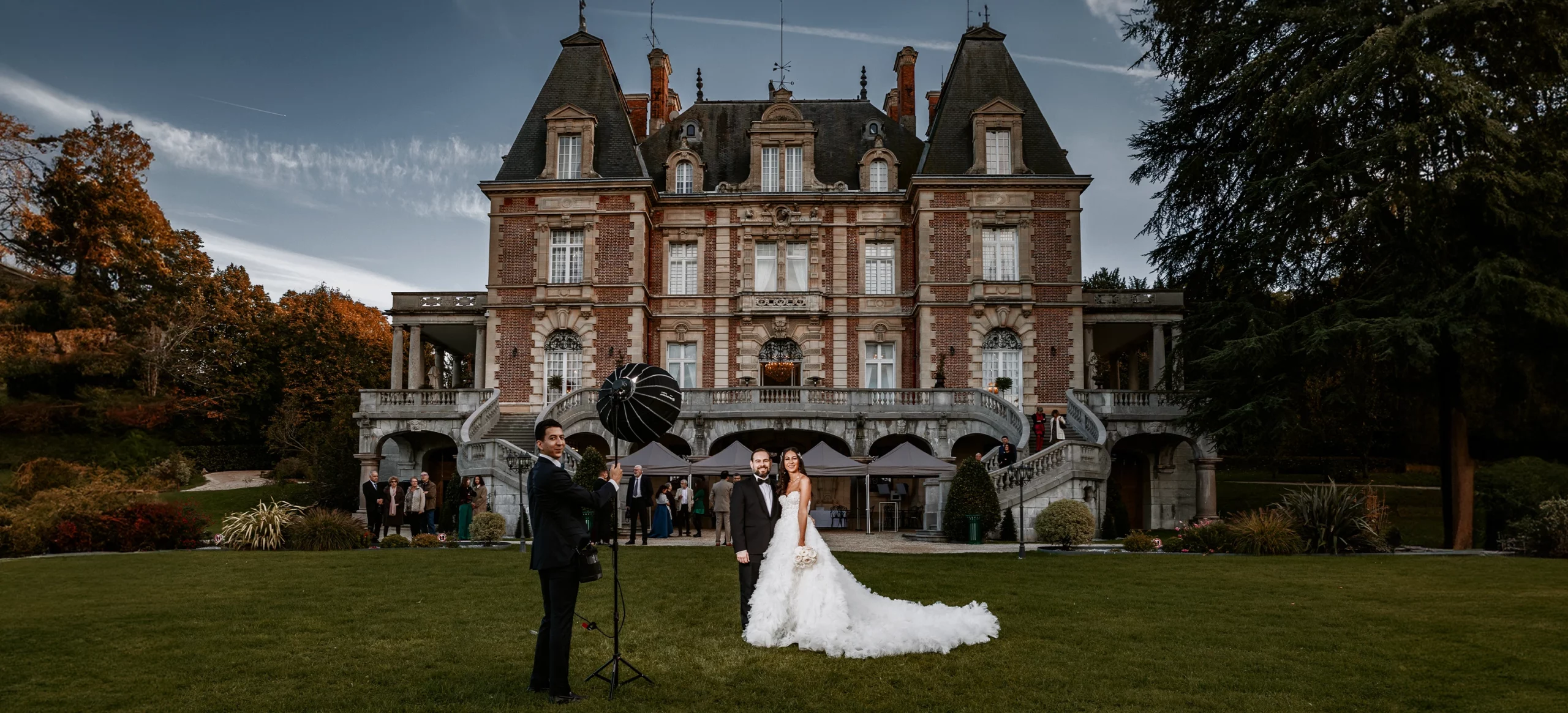 Vue large du Château Bouffémont avec les mariés posant devant, un jeune homme tenant un flash déporté Elinchrom sur le côté. Photo par Laurène Zabary