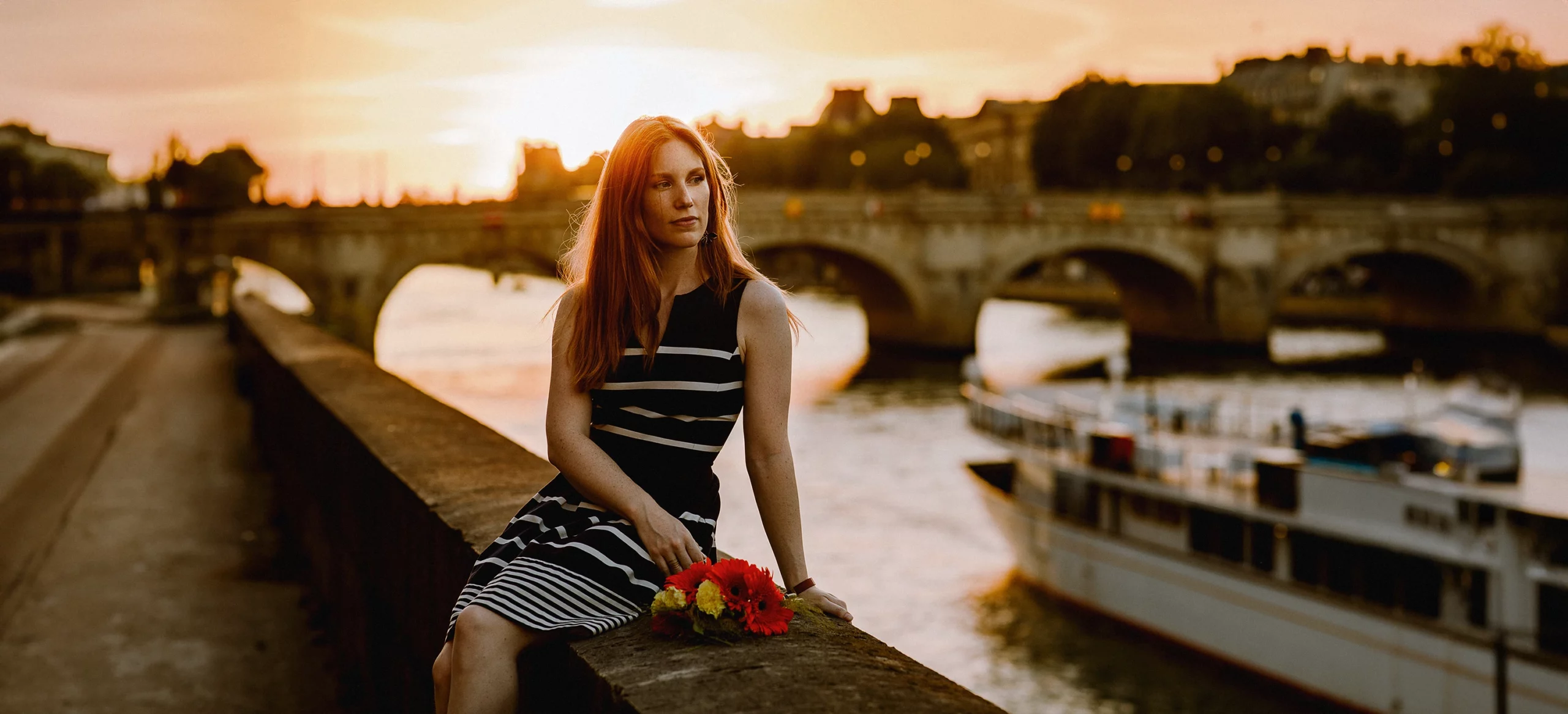 Fiona, jeune femme rousse, assise près de la Seine, un pont et une péniche en arrière-plan. Photo par Laurène Zabary