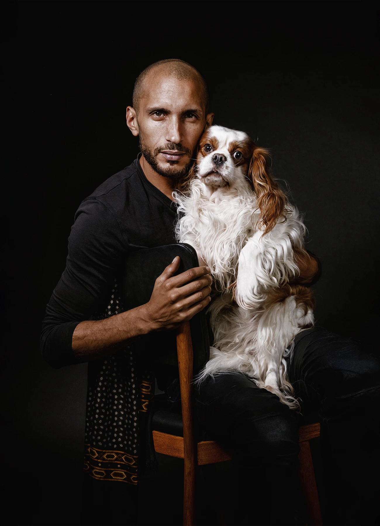Portrait de Mike, comédien, assis sur une chaise avec un chien sur ses genoux, sur fond noir.
