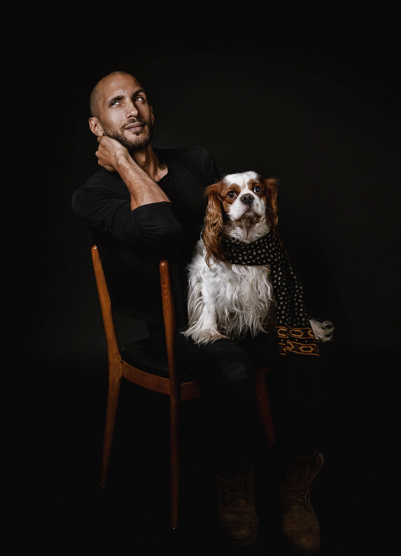 Portrait de Mike, comédien, assis sur une chaise avec un chien portant une écharpe sur ses genoux, sur fond noir.