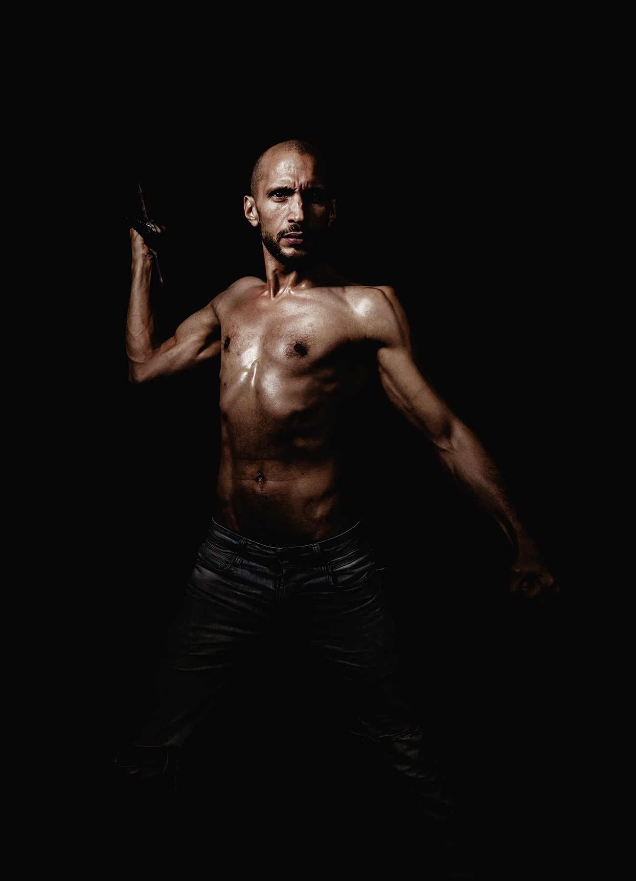 Comédien en position d’attaque, tenant une épée levée, éclairé par une lumière intense sur fond noir.