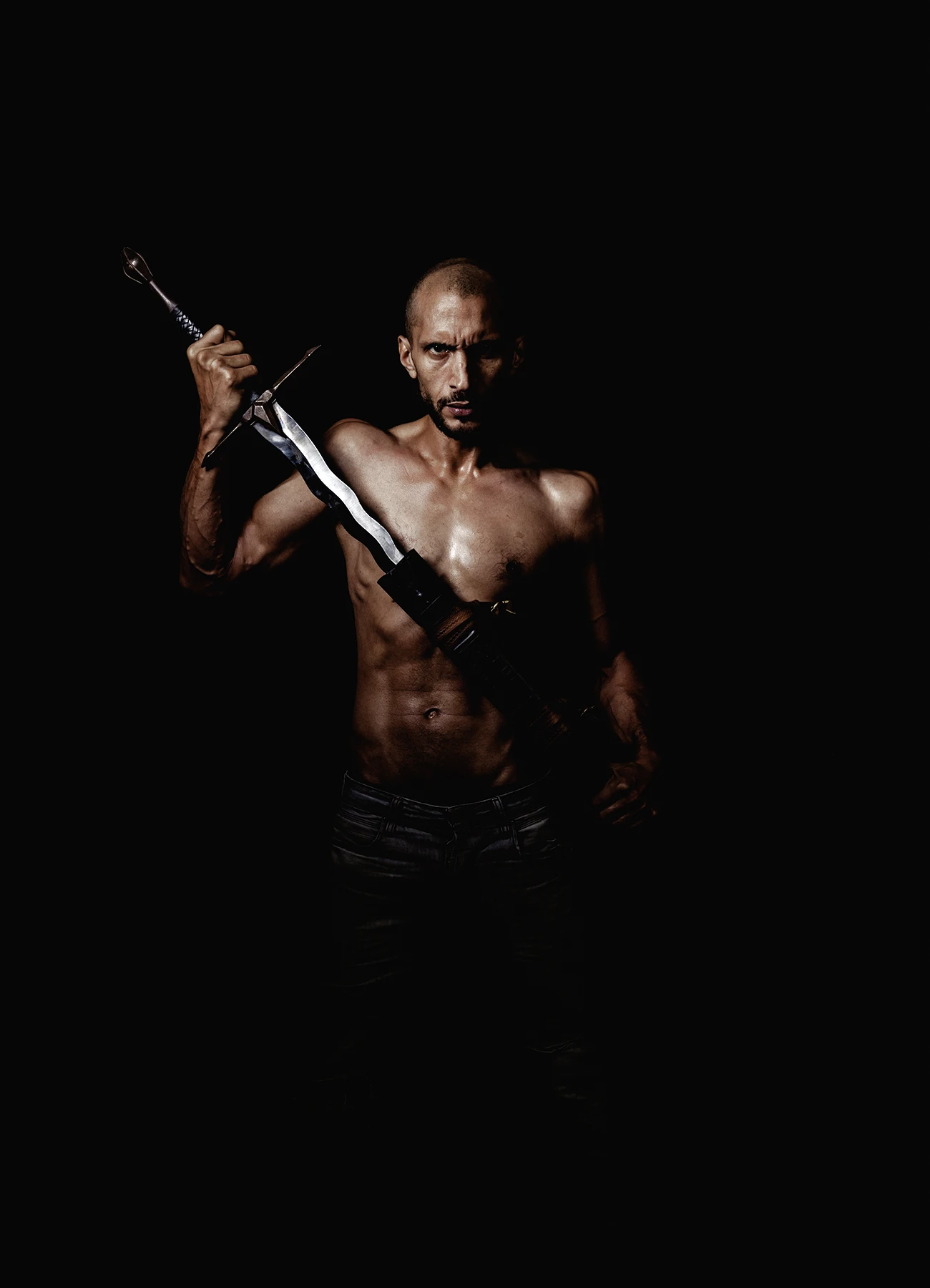 Portrait contrasté d’un comédien regardant fixement l’objectif, une épée à la main, sur fond noir.