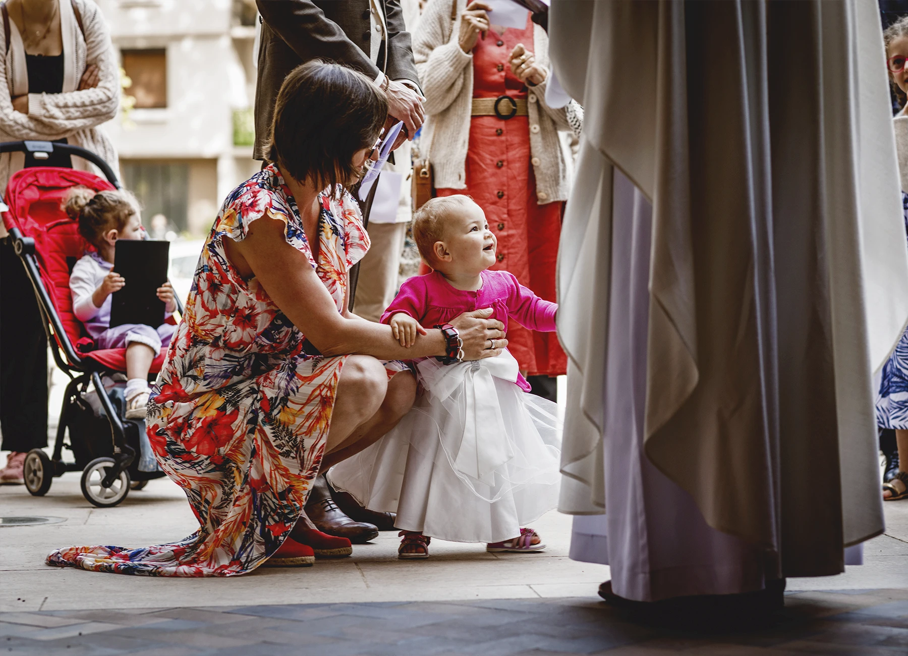 Une femme accroupie tient debout le bébé à baptiser entourée des membres de la famille dans l’église Saint-Joseph de Montrouge, photographiée par Laurène Zabary.