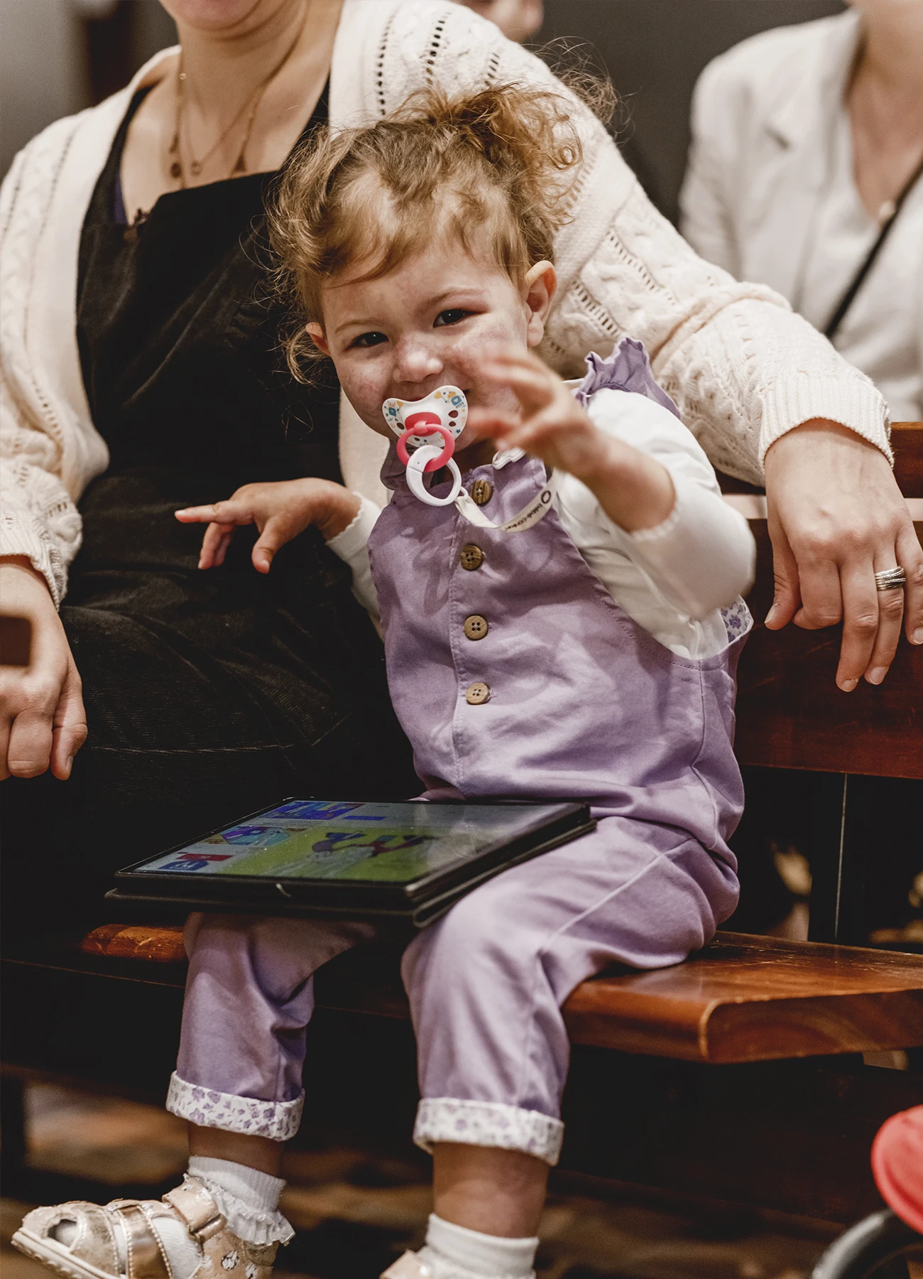 Une petite fille avec une tototte, tenant une tablette électronique sur les genoux, capturée lors d’un baptême à Saint-Joseph de Montrouge par Laurène Zabary.