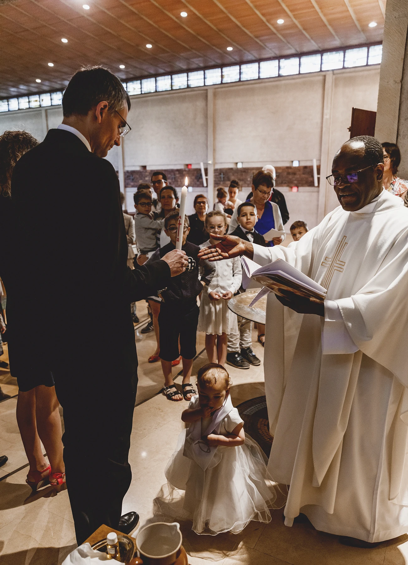 Le prêtre devant l’enfant et le témoin tenant un cierge lors d’un baptême à Saint-Joseph de Montrouge, capturé par Laurène Zabary.