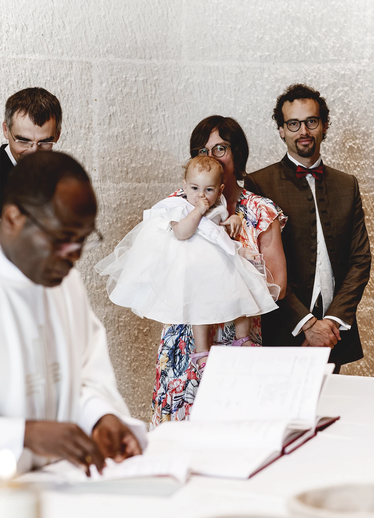 Le couple et le bébé à baptiser regardent la photographe pendant que le prêtre prépare les registres à l'église Saint Joseph de Montrouge, réalisé par Laurène Zabary, photographe à Paris.