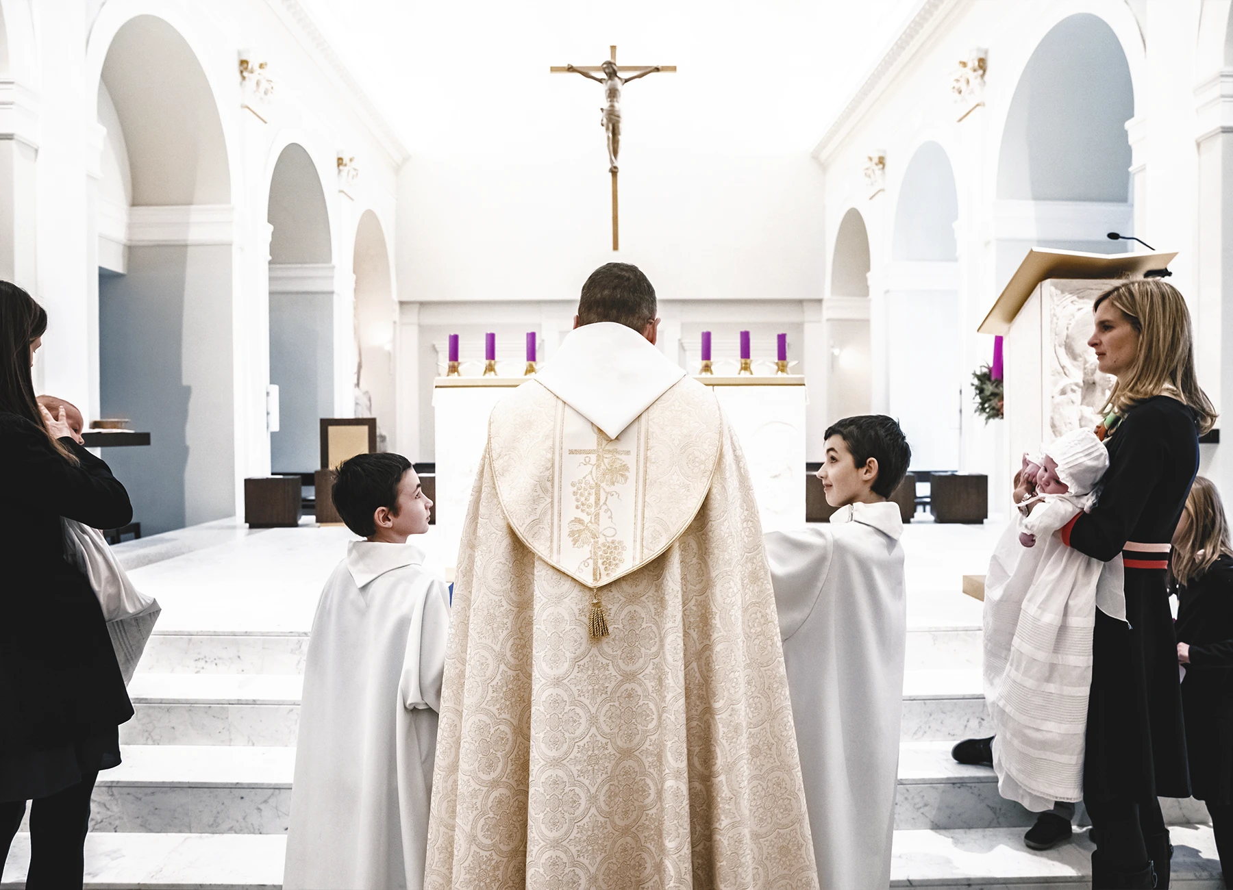 Le prêtre vu de dos, entouré de deux enfants de chœur, devant une croix.
