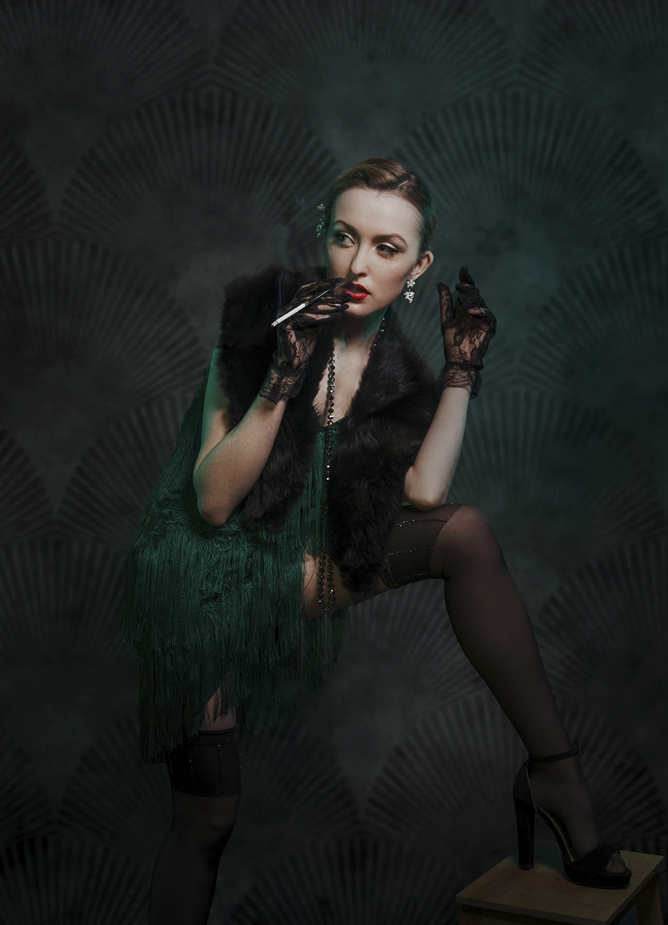 Kalysta en robe style années 20 et bas, porte un porte-cigarette à sa bouche avec une jambe posée sur un marchepieds, photo en studio réalisée par Laurène Zabary.