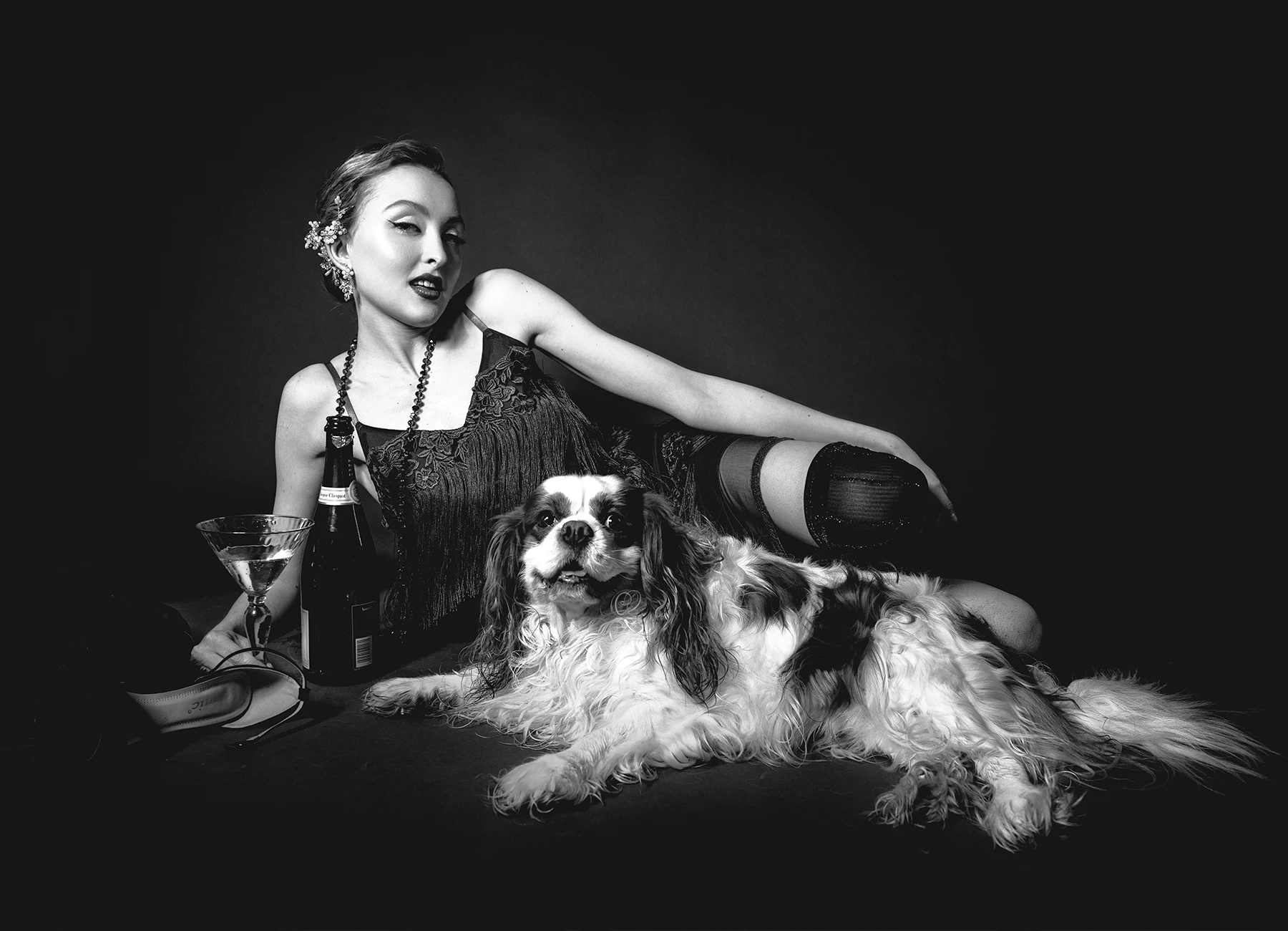 Kalysta en tenue style années 20, allongée avec une coupe et une bouteille de champagne, accompagnée d'un chien Cavalier King Charles, photo en studio réalisée par Laurène Zabary.