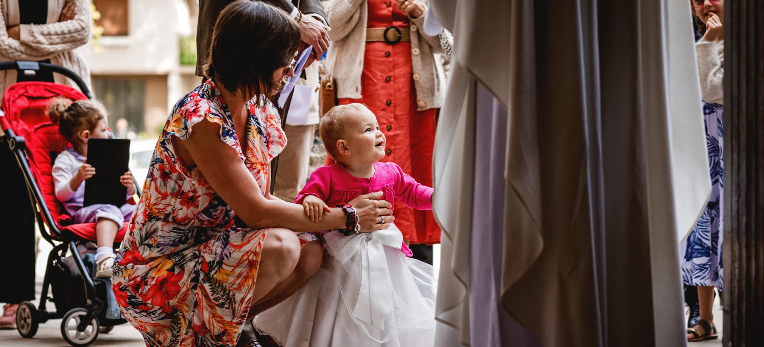 Une femme accroupie tient debout un bébé à baptiser, entourée des membres de la famille, lors d'un baptême à l'église Saint Joseph de Montrouge, réalisé par Laurène Zabary, photographe à Paris.