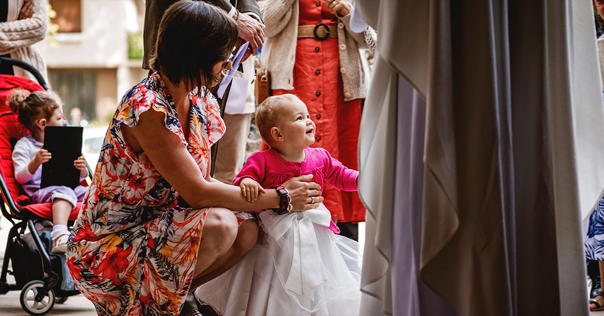 Une femme accroupie tient debout un bébé à baptiser, entourée des membres de la famille, lors d'un baptême à l'église Saint Joseph de Montrouge, réalisé par Laurène Zabary, photographe à Paris.