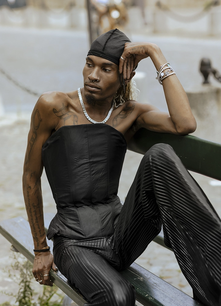 Be Norman pose sur un banc avec un corset noir et un pantalon plissé de sa marque Duhbuhlyoo