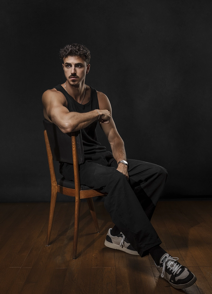 Portrait en pied de Noam assis sur une chaise dans une ambiance sombre, réalisé pour son book photo en studio à Paris